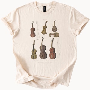 Può includere: T-shirt color crema con un'illustrazione vintage di vari strumenti a corda, tra cui violini e violoncelli, con notazione musicale. Il design presenta una varietà di forme e dimensioni degli strumenti, con un'estetica classica e antica.