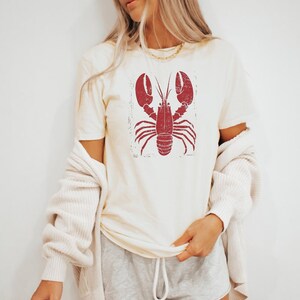 以下が含まれることがあります： クリーム色のTシャツに、赤いビンテージスタイルのロブスターのグラフィックがプリントされています