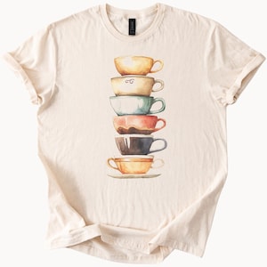May include: A cream-coloured t-shirt displaying a watercolour illustration of six stacked teacups in various colours. The cups are yellow, light brown, teal, red, dark brown, and orange. The shirt is made of soft material.