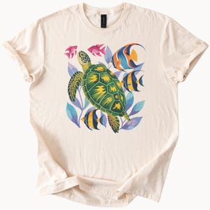 May include: Cream-coloured t-shirt with a colourful illustration of a green sea turtle with yellow details, surrounded by fish and blue and purple leaves. The design is centred on the front of the shirt.