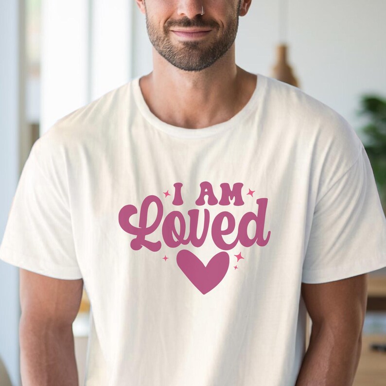 Puede incluir: Camiseta blanca con el texto "I AM Loved" en rosa, acompa&ntilde;ada de un coraz&oacute;n rosa. La camiseta est&aacute; hecha de un material suave, adecuada para uso casual.
