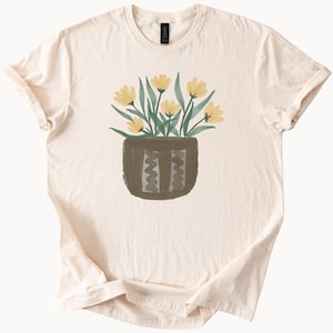 May include: Cream-colored t-shirt featuring a watercolor illustration of yellow flowers in a brown pot. The pot has a textured design. The shirt has short sleeves and a crew neck.