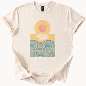 以下が含まれることがあります： クリーム色の半袖Tシャツで、中央にグラフィックプリントが施されています。デザインは、黄色の円の中にあるピンクの太陽と、その下の黄色の地平線、そして太陽の下に水を表す青と黄色の波線が描かれています。