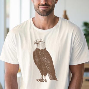 May include: A white t-shirt with a brown and white illustration of a bald eagle.