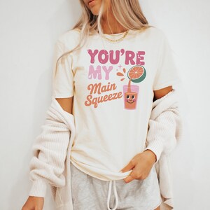 K&ouml;nnte beinhalten: Cremefarbenes T-Shirt mit dem Text "YOU'RE MY Main Squeeze" in rosa und orangefarbenen Buchstaben. Eine Grafik eines l&auml;chelnden Saftglases und einer halben Grapefruit ist ebenfalls abgebildet. Das Shirt ist ein l&auml;ssiges Kleidungsst&uuml;ck f&uuml;r den Alltag.