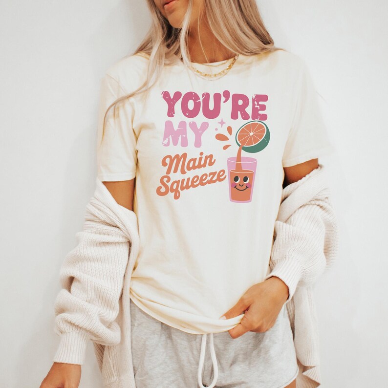 K&ouml;nnte beinhalten: Cremefarbenes T-Shirt mit dem Text "YOU'RE MY Main Squeeze" in rosa und orangefarbenen Buchstaben. Eine Grafik eines l&auml;chelnden Saftglases und einer halben Grapefruit ist ebenfalls abgebildet. Das Shirt ist ein l&auml;ssiges Kleidungsst&uuml;ck f&uuml;r den Alltag.