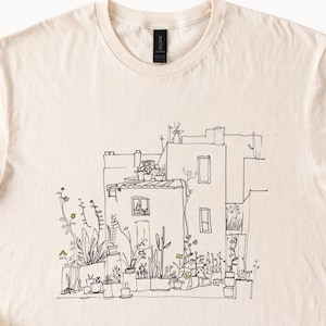 May include: Cream-colored t-shirt featuring a black and white line drawing of a building with potted plants and flowers. The illustration depicts a building with windows, a rooftop, and various plants.