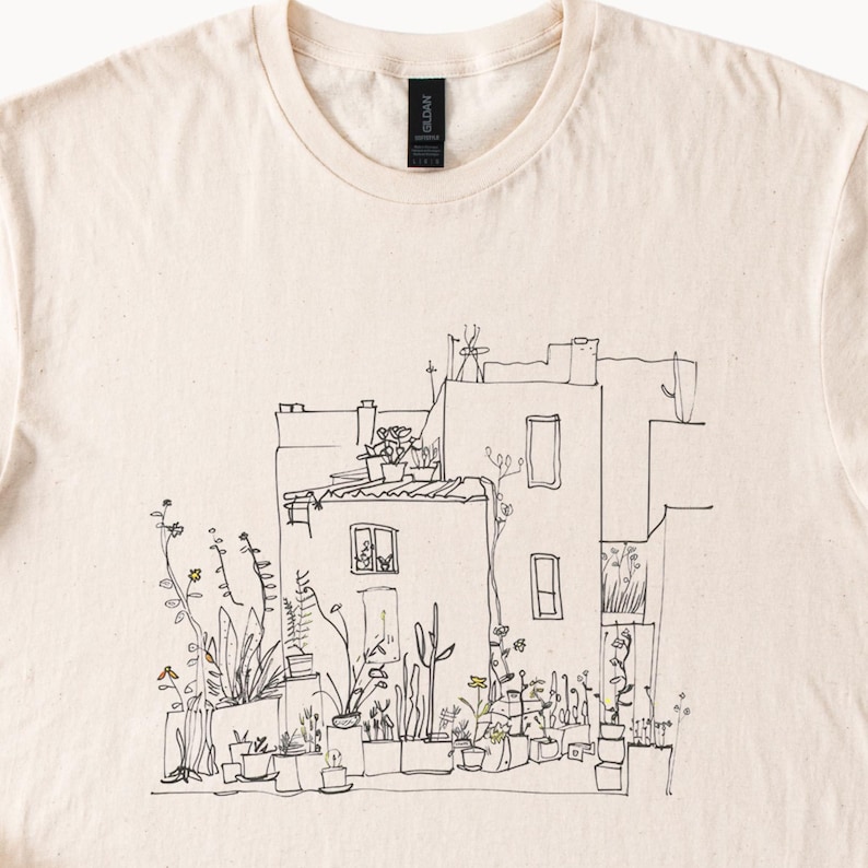 May include: Cream-colored t-shirt featuring a black and white line drawing of a building with potted plants and flowers. The illustration depicts a building with windows, a rooftop, and various plants.