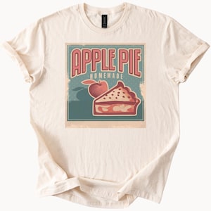 Puede incluir: Camiseta color crema con un diseño gráfico de estilo vintage. El diseño incluye las palabras "APPLE PIE HOMEMADE" en una fuente retro, junto con una ilustración de una manzana y una porción de tarta. El diseño está sobre un fondo verde azulado.