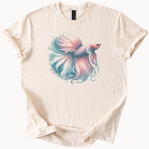 May include: Cream-colored t-shirt featuring a detailed illustration of a betta fish. The fish has a flowing tail in shades of pink and blue. The design is centered on the shirt, with the sleeves rolled up.