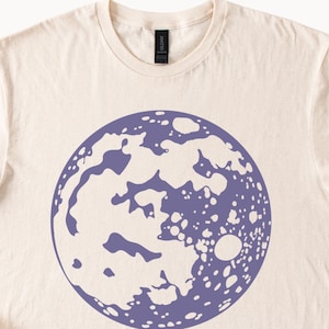 Op de afbeelding: Een licht beige t-shirt met een paarse maanprint. De maan is een gedetailleerde illustratie met kraters en een witte omtrek.