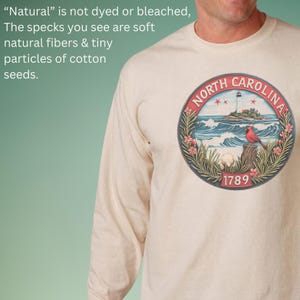 May include: Cream-colored long-sleeve shirt featuring a circular graphic of North Carolina's coastline, a lighthouse, and a red cardinal. The graphic is framed by a navy blue border with the text "NORTH CAROLINA" and the year "1789". The shirt is made of natural fibers.