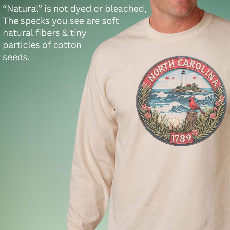May include: Cream-colored long-sleeve shirt featuring a circular graphic of North Carolina's coastline, a lighthouse, and a red cardinal. The graphic is framed by a navy blue border with the text "NORTH CAROLINA" and the year "1789". The shirt is made of natural fibers.