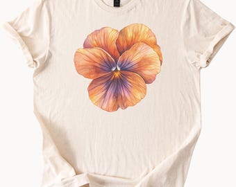 May include: Cream-colored t-shirt featuring a large, watercolor-style pansy flower. The flower has orange and purple petals with a yellow center. The shirt has short sleeves and a crew neck.