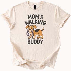 Puede incluir: Camiseta color crema con el texto "MOM'S WALKING BUDDY" arriba y abajo de un perro de dibujos animados. El perro es marrón y canela, sosteniendo una correa en la boca. Prenda de vestir informal.