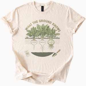 May include: A cream-colored t-shirt featuring a graphic of three root vegetables labeled Golden Beet, Purple Top, and Celeriac. The text WHAT THE GROUND KNOWS arches above the plants, with a small seed packet and a hand trowel illustrated below.