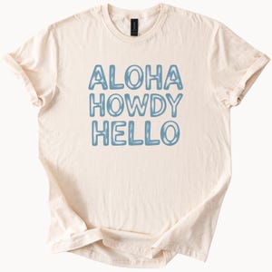 May include: Cream-colored t-shirt with the words "ALOHA HOWDY HELLO" in light blue, stacked vertically. The shirt has short sleeves and a crew neck. The fabric appears soft and slightly wrinkled.
