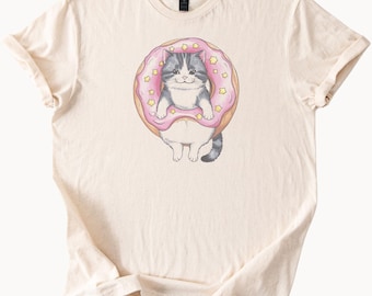 Könnte beinhalten: Cremefarbenes T-Shirt mit einer Cartoon-Katze in einem rosa Donut mit gelben Sternstreuseln. Die Katze hat grau-weißes Fell und lächelt. Das Shirt hat einen Rundhalsausschnitt und kurze Ärmel.