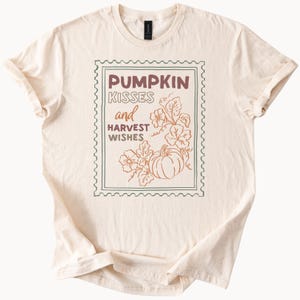 Peut inclure: T-shirt crème avec un motif rectangulaire marron et vert. Le motif comprend les mots "PUMPKIN KISSES and HARVEST WISHES" et une illustration de citrouille et de feuilles. Le t-shirt a des manches courtes et un col rond.