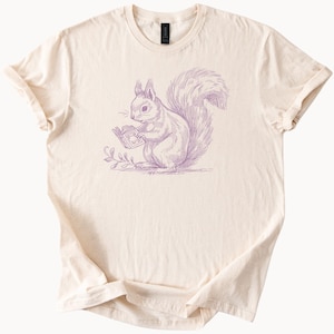 May include: Cream-colored t-shirt featuring a purple illustration of a squirrel reading a book. The squirrel is depicted in a detailed, vintage style, holding a small book and sitting near a sprig of leaves. The shirt has a classic crew neck and short sleeves.