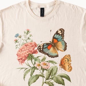 May include: Cream-colored t-shirt featuring a detailed illustration of two butterflies and floral elements. The butterflies have orange, blue, and brown wings. The flowers are pink, with green leaves and stems. The shirt has a classic crew neck.