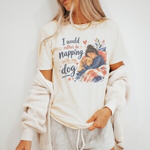 May include: A cream-colored short-sleeve t-shirt featuring a graphic illustration of a person cuddling a golden dog under a blanket. The text reads, I would rather be napping with my dog, accompanied by small red hearts and blue floral accents.