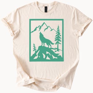 May include: Cream-colored t-shirt featuring a teal graphic of a howling wolf on a mountain, framed by a rectangular border. The design includes mountains and pine trees, creating a nature-inspired theme.