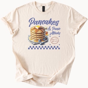 Könnte beinhalten: Ein beiges T-Shirt mit einem Grafikdruck von einem Stapel Pfannkuchen mit Blaubeeren und Sirup. Der Text "Pancakes & Panic Attacks" befindet sich über den Pfannkuchen und "Buy one get one" darunter.