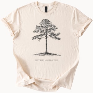 Puede incluir: Camiseta color crema con una ilustración detallada en blanco y negro de un pino de hoja larga del sur. El texto "SOUTHERN LONGLEAF PINE" está impreso debajo. La camiseta tiene cuello redondo y mangas cortas.