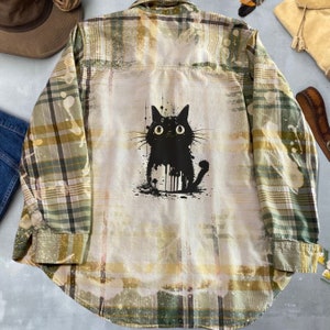 Puede incluir: Una camisa de franela a cuadros verde y amarilla con un gráfico de gato negro en la espalda. El gato está en un estilo de pintura salpicada.