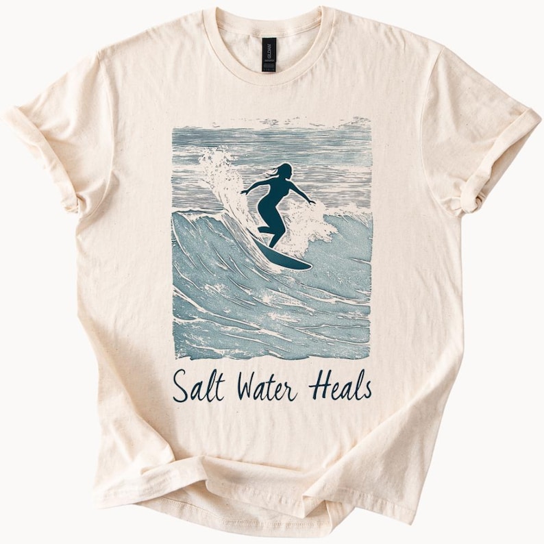 May include: A beige t-shirt with a blue and white illustration of a person surfing on a wave. The text "Salt Water Heals" is printed below the illustration.