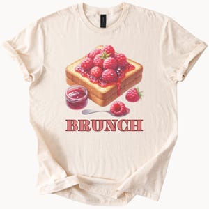 May include: A white t-shirt with a graphic of a slice of toast topped with raspberry jam and fresh raspberries. The text "BRUNCH" is printed below the toast.