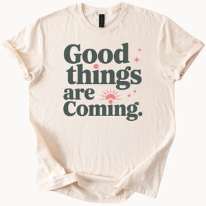 May include: A cream-coloured t-shirt with the text "Good things are coming." in dark green letters. A pink sun with a red circle in the centre is above the word "coming".