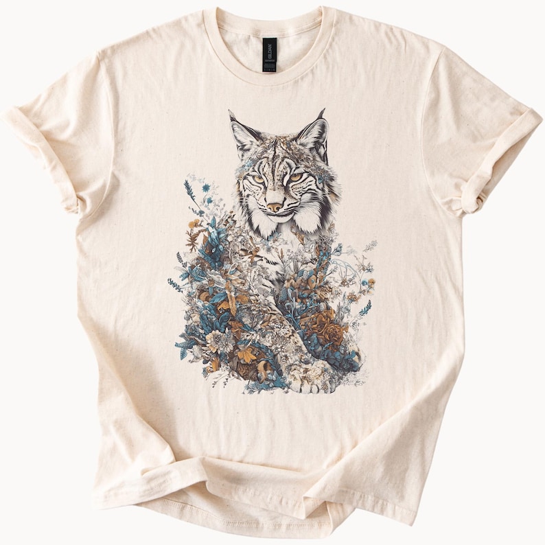 May include: Cream-colored t-shirt featuring a detailed illustration of a lynx surrounded by flowers and foliage in shades of blue, brown, and green. The graphic is centered on the front of the shirt, creating a nature-inspired design.