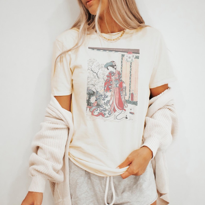 May include: Cream-colored t-shirt featuring a Japanese woodblock print of a woman in a red kimono. The print includes a detailed depiction of a traditional scene with floral and architectural elements. The shirt has short sleeves and a relaxed fit.