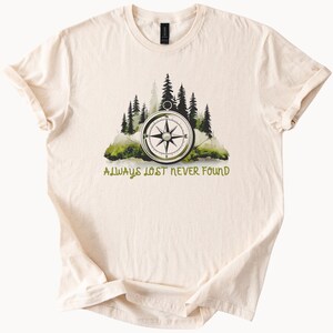 Può includere: Una t-shirt color crema con un design a bussola verde e marrone e il testo "Always Lost Never Found".