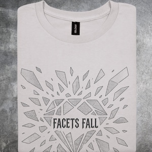 May include: A light gray crew neck t-shirt featuring a black line art graphic of a diamond shattering into multiple shards. The text FACETS FALL is printed in a bold, stylized font within the center of the diamond design on the front of the shirt.
