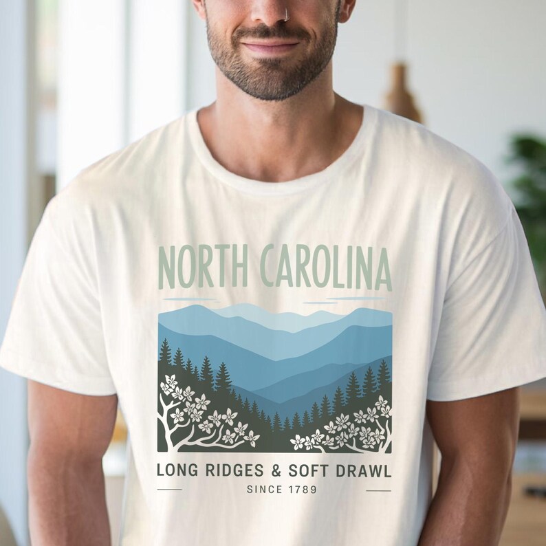 May include: White t-shirt featuring a landscape design with the text "NORTH CAROLINA" above a mountain scene in shades of blue and green. Below the image is the text "LONG RIDGES & SOFT DRAWL" and "SINCE 1789".