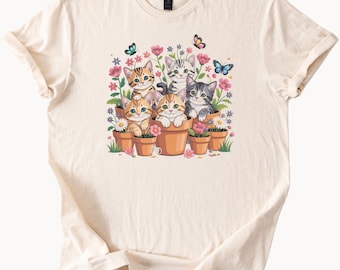 May include: Cream-colored t-shirt featuring a colorful graphic of six kittens in terracotta flower pots surrounded by pink flowers and butterflies. The kittens have various tabby patterns and colors. The shirt has a crew neck and short sleeves.