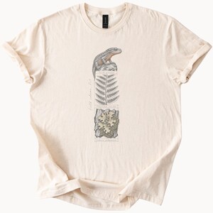 May include: A cream-colored t-shirt featuring a vertical graphic illustration of a lizard perched on a rock, a fern leaf, and a patch of lichen. The design includes the text "field column, Oct" and scientific labels in a thin black font.