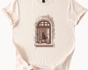 May include: A cream colored t-shirt with a black cat sitting in a window with potted plants. The window is arched and has a brown frame. The cat is looking out the window.