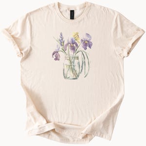 May include: Cream-colored t-shirt featuring a watercolor illustration of purple and yellow irises in a clear glass jar. The floral design includes green leaves and stems. The shirt has a crew neck and short sleeves.