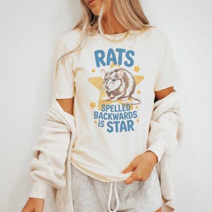 Puede incluir: Camiseta color crema con el texto azul "RATS SPELLED BACKWARDS IS STAR". El gráfico presenta una rata marrón y blanca sobre una estrella amarilla con estrellas amarillas más pequeñas. Prenda de vestir informal.