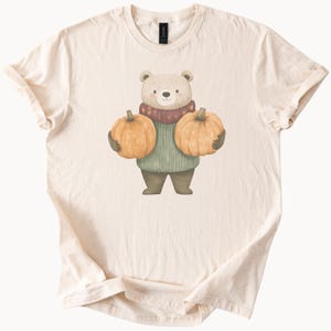 Könnte beinhalten: Cremefarbenes T-Shirt mit einem Cartoon-Bären, der zwei orangefarbene Kürbisse hält. Der Bär trägt einen grünen Pullover und einen Schal mit roten und lila Akzenten. Das Design ist im Aquarellstil gehalten, ideal für den Herbst.