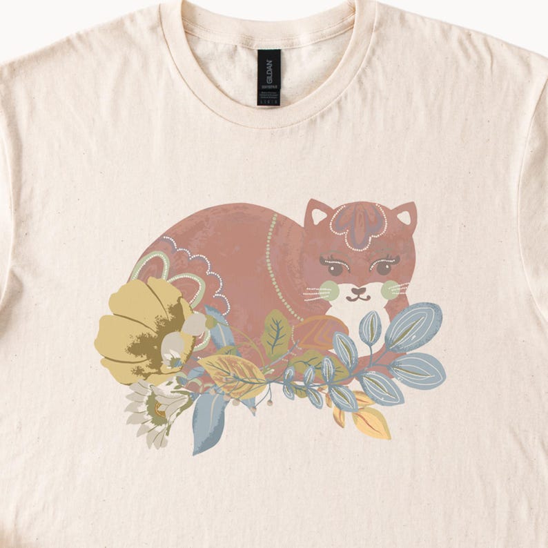 May include: A cream-coloured t-shirt with a cat illustration and floral details. The cat is in shades of brown and pink, with yellow, blue, and green leaves and flowers. The design is centrally placed on the shirt, offering a whimsical aesthetic.