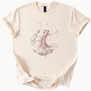 Puede incluir: Camiseta color crema con una ilustración enmarcada de una figura bailando con un vestido fluido. La bailarina está representada en tonos morados y rosas, con tela ondeando a su alrededor. La camiseta tiene cuello redondo y mangas cortas.