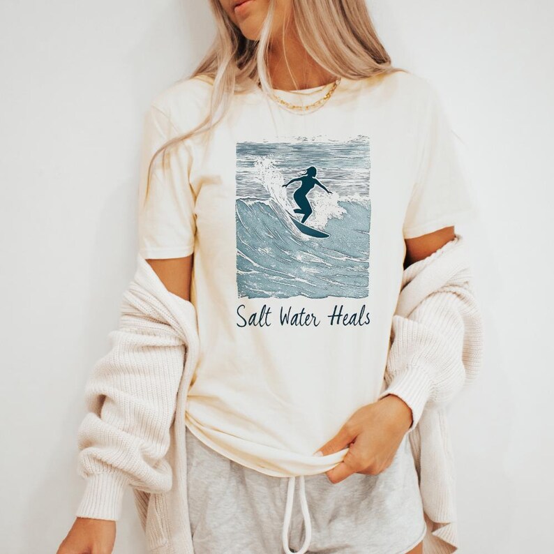 May include: A cream colored t-shirt with a graphic of a person surfing on a wave. The text "Salt Water Heals" is printed below the graphic.