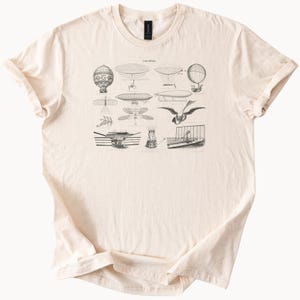 May include: Cream-colored t-shirt featuring a detailed black and white illustration of various vintage airships and flying machines. The design includes hot air balloons, dirigibles, and early aircraft, creating a historical and scientific theme.