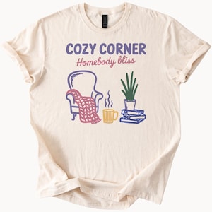 May include: Cream-colored t-shirt with a graphic design featuring a cozy scene. The design includes a chair with a blanket, a mug, a plant, and books. Text above the image reads "COZY CORNER Homebody bliss."