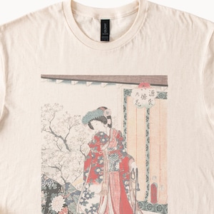 May include: Cream-colored t-shirt featuring a Japanese woodblock print. The print depicts a woman in a red kimono with a floral design, standing near a building with Japanese text. The shirt has a crew neck and short sleeves.